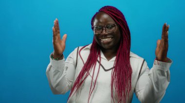 Mavi arka planda örgülü saçları olan, gözlüklü ve beyaz gömlekli neşeli ifadesini ve canlı tarzını vurgulayan bir kadın..