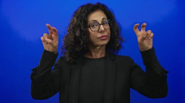 Orta yaşlı İspanyol bir kadın, parmaklarıyla tırnak işaretleri yaparak izole edilmiş mavi bir arkaplanı işaret ediyor, özgüven ve ifade yayıyor..