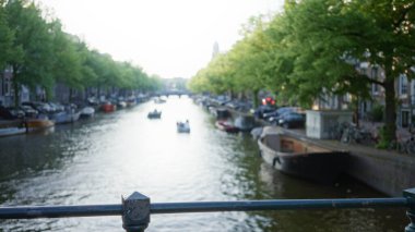 Amsterdam 'daki bulanık kanal sahnesi. Suda yüzen tekneler, kıyıları kaplayan ağaçlar ve arka planda çözünmüş şehir manzarası. Huzurlu bir Alman atmosferi yaratıyor..