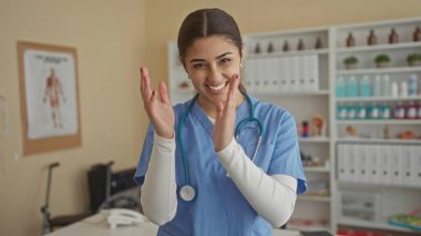 İspanyol üniformalı kadın hemşire hastane odasında sevinçle gülümsüyor. Arka planda tıbbi ekipmanlar var. Sıcaklık ve profesyonelliği yansıtıyor..