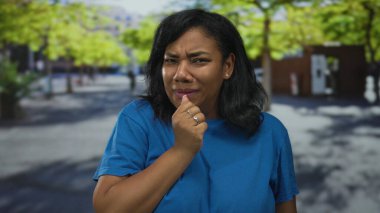 Tuğla bina ve park edilmiş arabaların yakınındaki Sunlit City Caddesi 'nde düşünceli bir hareketle çeneye dokunan kadın..