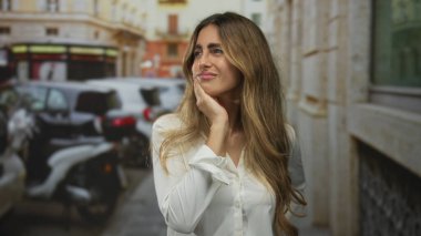 Beyaz bluzlu, sarı saçlı bir kadın iki eliyle yanağını dar bir caddede tutuyor; diş ağrısı rahatsızlığı..