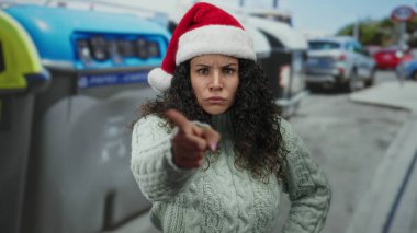 Noel Baba şapkalı İspanyol kadın, arka planda arabaları ve geri dönüşüm kutularıyla şehir sokağında, neşeli ve iddialı bir açık hava sahnesini yakalıyor..