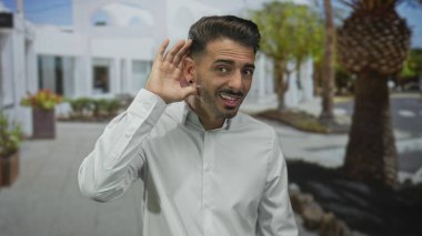 Beyaz binalar ve palmiye gövdeleri ile çevrili güneşli bir caddede eli kulağında bir adam; merak.