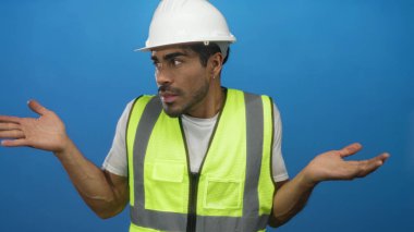 Çağdaş stüdyoda çelik yelek ve sert şapkalı bir adam avuçlarını mavi zemine yaslıyor; belirsizlik.