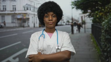 Steteskoplu kadın doktor, beyaz binalarla dolu bir caddede dururken çenesini uzatıyor ve kollarını kavuşturuyor..