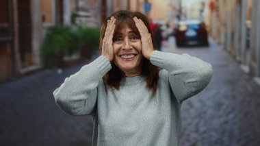 Yaşlı İspanyol kadın şehir sokaklarında kulaklarını tıkıyor, stres veya hayal kırıklığını dile getiriyor, gri bir kazak giyiyor, şehir hayatını ve duygularını yakalıyor..