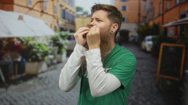 Gönüllü gömleği olan sakallı bir adam kaldırım taşı sokağındaki kameraya öpücük hareketi yapıyor..