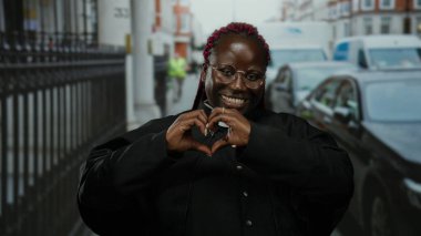 Papaz gibi giyinmiş, gülümseyen ve kalbi şekillendiren bir kadın kalabalık bir şehir caddesinde, arka planda arabalar ve binalarla, dışarıda neşe ve pozitifliği ifade ediyor..