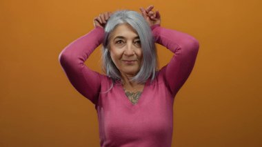 Gri saçlı yaşlı kadın, elleri turuncu arka planda, pembe bir kazak giyiyor, mizahi ve neşeli bir sahne yaratıyor..