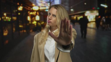 Sarışın bir kadın, yoğun bir şehir caddesinde gece bir durak hareketi yapıyor. Açık hava ortamında kendine güveni ve kararlılığı iletiyor..