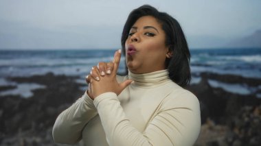 Sahilde parmak tabancasıyla jest yapan bir kadın, kendinden emin bir ifadeyle neşeli bir anı yakalıyor, çerçevedeki güzelliği ve doğayı vurguluyor..