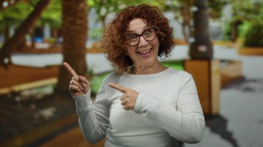 Parkta, yeşil yeşil bir ortamda gülümseyen, sıradan beyaz kıyafetler ve gözlük takan, neşe ve mutluluk saçan bir kadın..