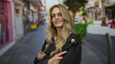 Sarışın genç bir kadın yargıç açık havada, hareketli bir sokakta elinde tokmakla duruyor ve yasal otoriteyi şehir hayatıyla harmanlıyor..