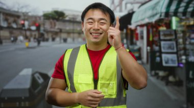 Şehir sokağında arka planda dükkanları olan düşünceli bir jest yapan yansıtıcı yelek giymiş bir adam..