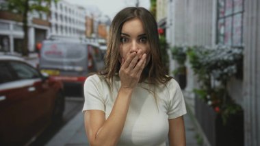 Beyaz tişörtlü, ağzı kalabalık şehir caddesinde park halindeki araba ve binalarla dolu genç İspanyol bir kadın şaşkınlık içinde şaşkınlık içinde..