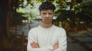 Beyaz süveter giyen ve gözlüklü bir adam ağaçlarla ve patikayla kaplı yeşil parkta kollarını kavuşturuyor..