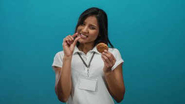 Meraklı yüzü olan genç bir kadın mavi arka planda küçük bir kurabiye tutuyor, beyaz gömlek ve isim kartı takıyor, parmakları olan küçük bir şey öneriyor..