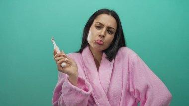 Pembe bornozlu kadın elinde elektrikli diş fırçasıyla yeşil duvara yaslanmış güvenli bir şekilde poz veriyor ve neşeli tavırlarını sergiliyor.