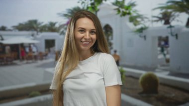 Güneşli bir avluda, yeşil ve beyaz mimarisi huzurlu bir ortamda gülümseyen bir kadın..