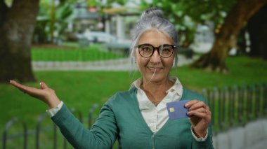 Parktaki son sınıf öğrencisi kadın elinde kredi kartıyla güneşli bir günde açık havada neşe saçıyor, gözlük takıyor ve yeşil hırka giyiyor..