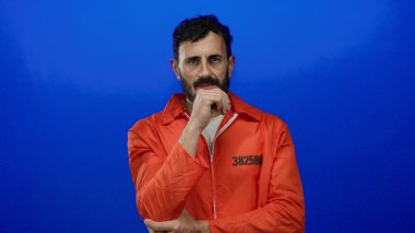 Turuncu hapishane tulumu giyen İspanyol sakallı adam mavi arka planda duruyor, eli çenesinde dalgın görünüyor..