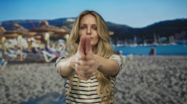 Kadın elini kumsala doğru uzatarak güneşli gökyüzünün altında tatil yaparken deniz kenarında genç bir sarışın havası yakalıyor..