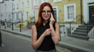 Şehir caddesinde telefonla konuşurken gülümseyen, gözlüklü ve siyah bluzlu, renkli binalarla çevrili, mutluluk ve kendine güvenini ifade eden bir kadın..