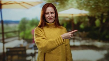 Şehir terasında sarı kazaklı, kırmızı saçlı bir kadın açık hava ortamında yoğun görünüyor, bulanık yeşillik ve veranda şemsiyeleriyle çevrili, düşünceli bir anı yakalıyor..