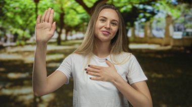 Genç bir kadın, canlı yeşil ağaçlarla çevrili güneşli bir parkta yemin ediyor, samimi bir ifadeyle, elini kaldırıyor ve kalbini tutuyor, bir anlık sadakati somutlaştırıyor.