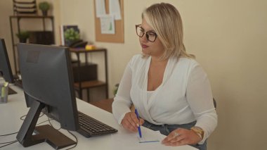 Kadın, kağıt notlar gösteren bilgisayar ve rafların yanındaki büro masasında kalemle yazıyor; konsantrasyon.