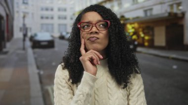 Kadın, park halindeki arabalar ve taş bina cepheleriyle dolu işlek şehir caddesinde parmaklarıyla yanağa dokunuyor..