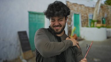 Güneşli bir günde İspanyol bir adam kırsal bir zemine karşı elinde bir panoyla omzunu tutarak rahatsızlık ya da yorgunluk hissi veriyor..