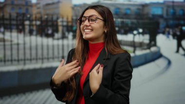 Kalabalık bir caddede neşeli bir şekilde el kol hareketi yapan, gözlüklü ve kırmızı bluzlu bir kadın, şehir enerjisiyle çevrili samimi ve canlı bir anı yakalıyor..