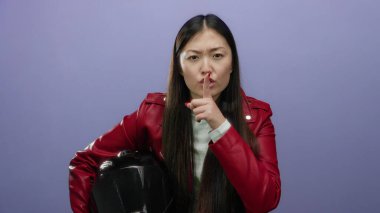 Kırmızı ceketli kadın miğfer tutarken mor arka planda sessizliği öneriyor. Dudaklarında parmaklarıyla poz veren genç Asyalı bir kadın var..