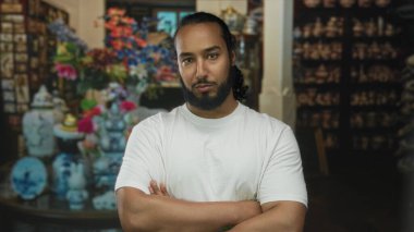 Beyaz tişörtlü ve sakallı, kolları kavuşturulmuş bir adam, sessiz bir kararlılık..