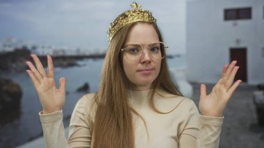 Altın tacı olan genç beyaz kadın deniz kenarındaki kıyıda omuz silkme hareketi yaparak el kaldırıyor..