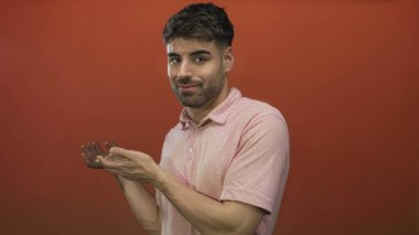 Portakal rengi stüdyo resmiyle ellerini sıvazlayan genç İspanyol adam pembe polo gömlek giyiyor ve nazik bir gülümseme sergiliyor..