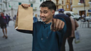 Sokak ortasında elinde kese kâğıdıyla gezen genç İspanyol adam gülümsüyor ve etrafı hareketli şehir manzaralarıyla çevrili..