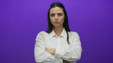 Beyaz gömlekli bir kadın, parlak mor bir duvarın önünde kolları çapraz duruyor, ciddi bir ifadeyi yansıtıyor, minimalist bir ortamda kararlılık ve güven öneriyor..