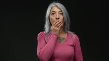 Pembe tişörtlü, gri saçlı yaşlı bir kadın siyah bir arkaplan karşısında el üstünde ağzıyla şok olmuş bir şekilde duruyor. Bir stüdyo çekiminde sürpriz ve duygu aktarıyor..