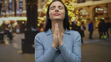 Üzgün ifadeli ve elleri olan genç bir kadın, arka planda şenlik ışıkları olan kalabalık bir caddede gece yalvaran bir jestte sıkıca tutundu..
