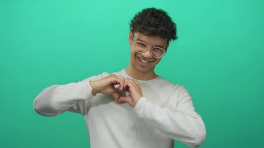 Gözlüklü, beyaz tişörtlü, parmak gösteren ve stüdyoda el kalbi şekli oluşturan bir adam..