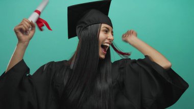 Diplomasını yeşil duvarda tutan akademik elbiseyle mezuniyeti kutlayan genç bir kadın..