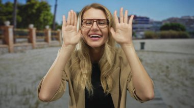 Güneşli bir parkta gülümseyen bir kadın, yüzünü açan eller, sarı saçlar, gözlük takan, açık yeşil bir ortamda neşe saçan bir kadın..