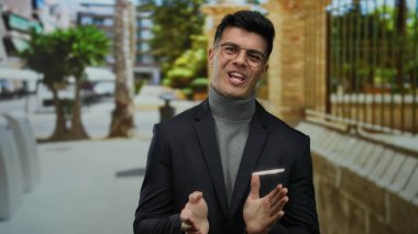 Gözlüklü genç İspanyol adam, bir şehir caddesinde gülümsüyor ve alkışlıyor, balıkçı yaka ve ceket giyiyor güneşli bir şehir zeminine karşı.