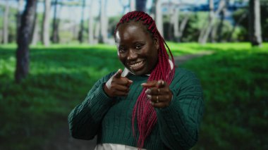 Pembe saçlı, gülümseyen ve açık hava parkında yeşil bir kazak giyen, mutluluk ve güven saçan Afrikalı bir kadın..