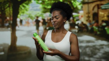 Güneşli şehir caddesinde elinde yeşil losyon şişesiyle gülümseyen bir kadın..