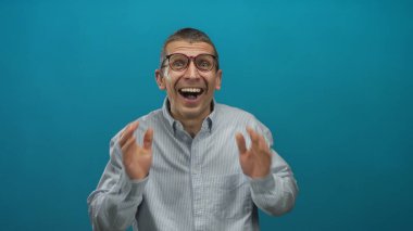 Mavi arka planda sürpriz yapan, beyaz gömlek ve gözlük takan, duygularını canlı bir şekilde sergileyen bir adam..