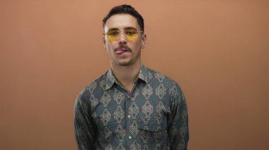 Güneş gözlüklü, desenli tişörtlü genç İspanyol bir adam kahverengi duvarın arkasına komik bir şekilde dil uzatıyor, izole bir çerçevede, tavır ve stil ifade ediyor..